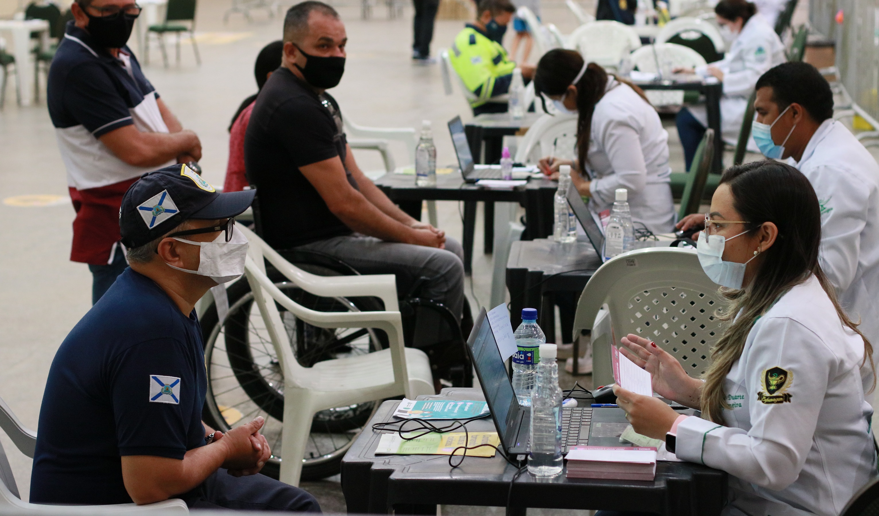 interior do centro de eventos, mostrando as pessoas sendo atendidas por profissionais da saúde em mesas, onde eles colhem os dados das pessoas que serão vacinadas